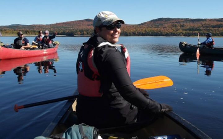 From the back of a canoe, the person in front turns around to smile at the camera. There are additional canoes floating on calm water in the background. 